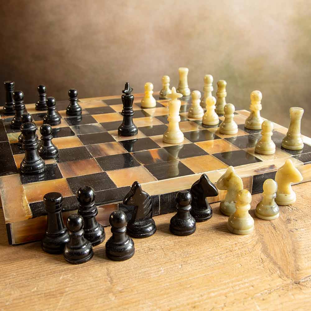 A polished chessboard with black and cream-coloured chess pieces arranged on a wooden table against a warm, blurred background.