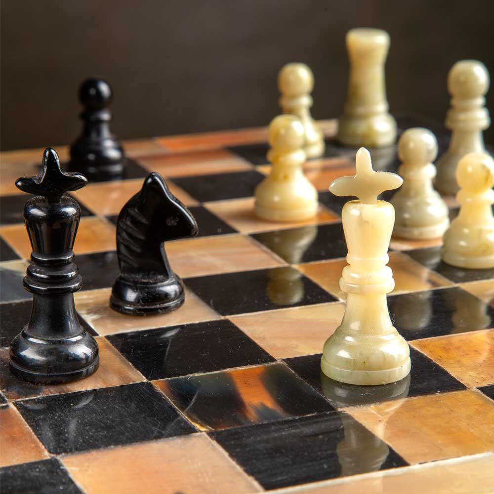 A close-up of a polished horn chessboard with alternating black and warm brown squares, featuring several glossy black chess pieces and cream-coloured pieces in the foreground and background.
