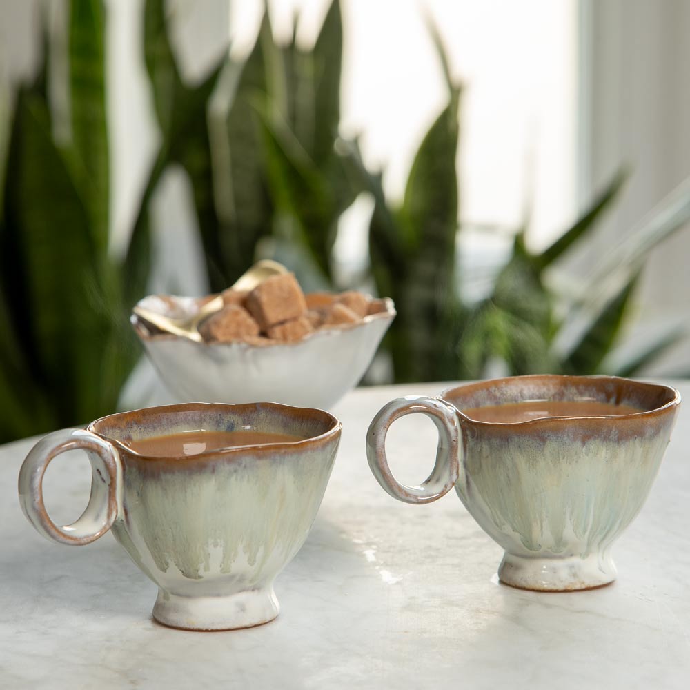Two ceramic cups with a light green and white textured glaze and a brown rim, filled with Hot Tea on a marble table with a bowl of sugar in the background.