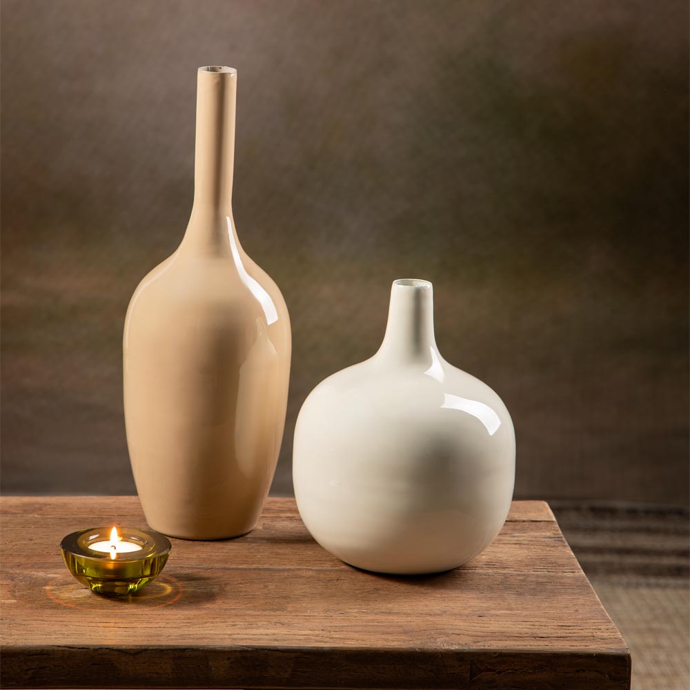 Two metal vases on a wooden table, one tall and beige with a narrow neck, and one short and white with a rounded bulbous shape, with a small lit candle in a green glass holder beside them.