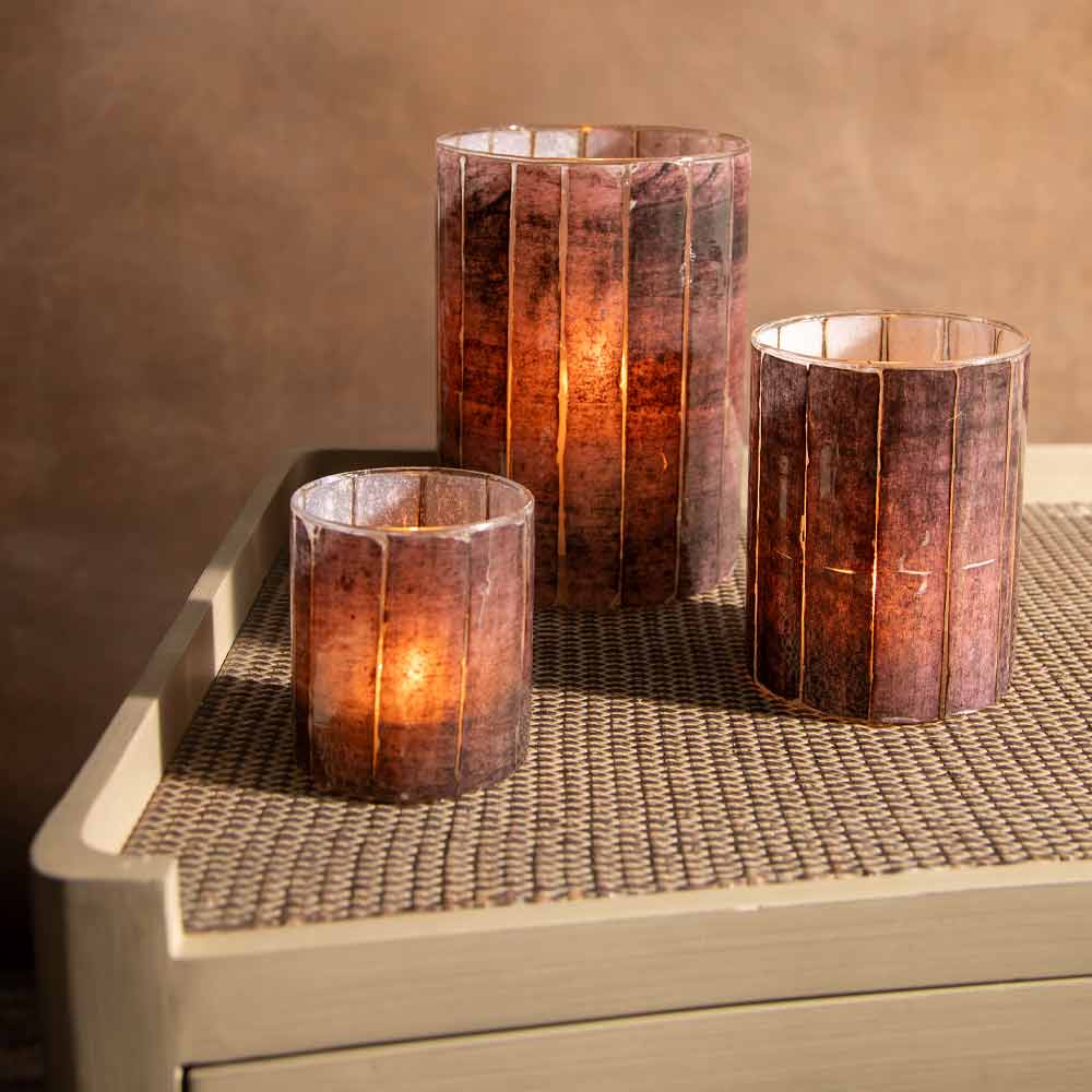 Three striped glass candle holders with a warm burgundy metallic finish, each containing a glowing candle, arranged on a light wood side table against a textured brown wall.