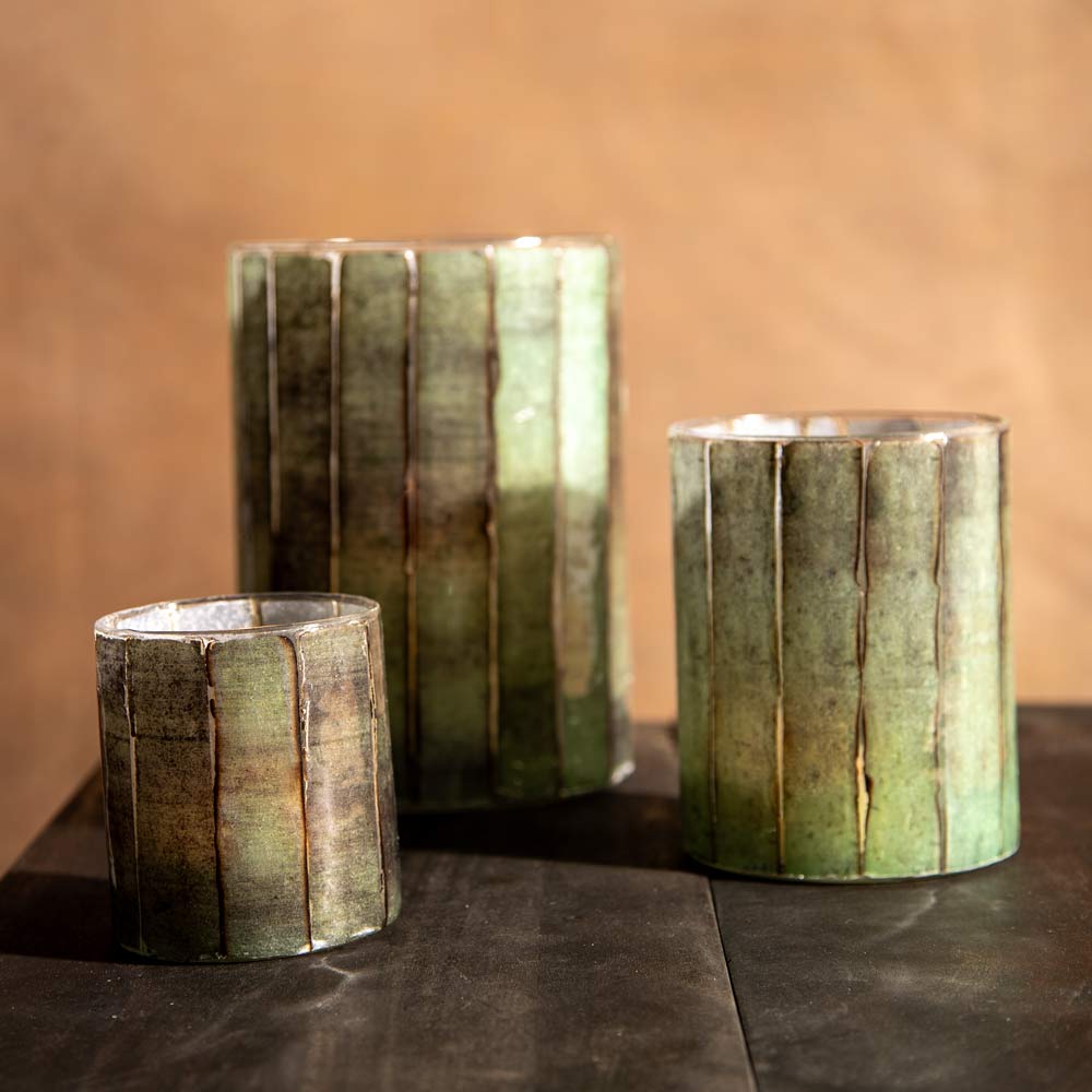 Three cylindrical candle holders with a textured, vertical ribbed design in olive green and brown tones, arranged on a dark tabletop against a warm brown background.