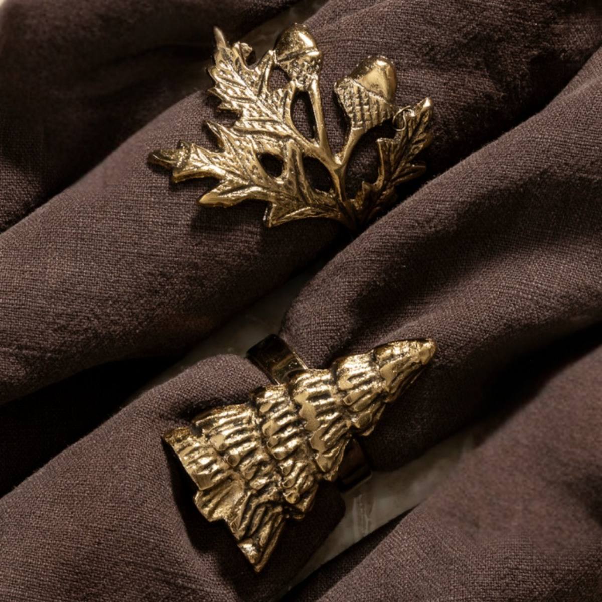 Joa Antique Gold Fir Tree and Gold Oak Leaf Napkin Rings on a brown fabric surface