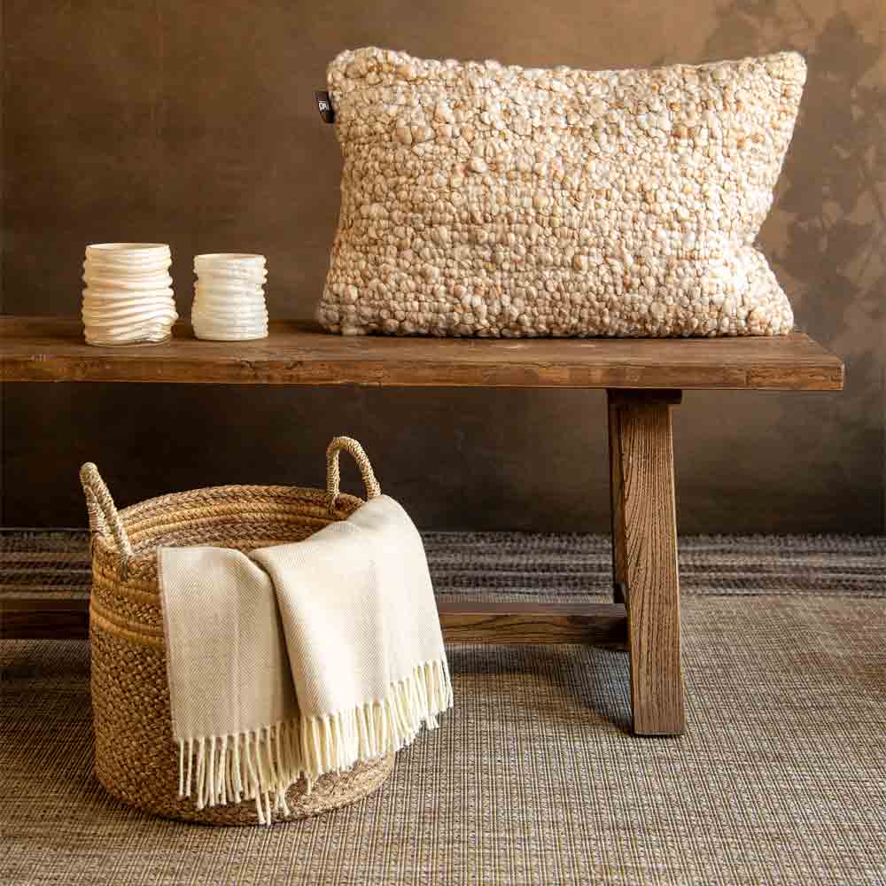 A textured cream bouclé style pillow displayed on a rustic wooden bench, with a woven basket holding a cream fringed throw blanket in the foreground against a warm brown wall.