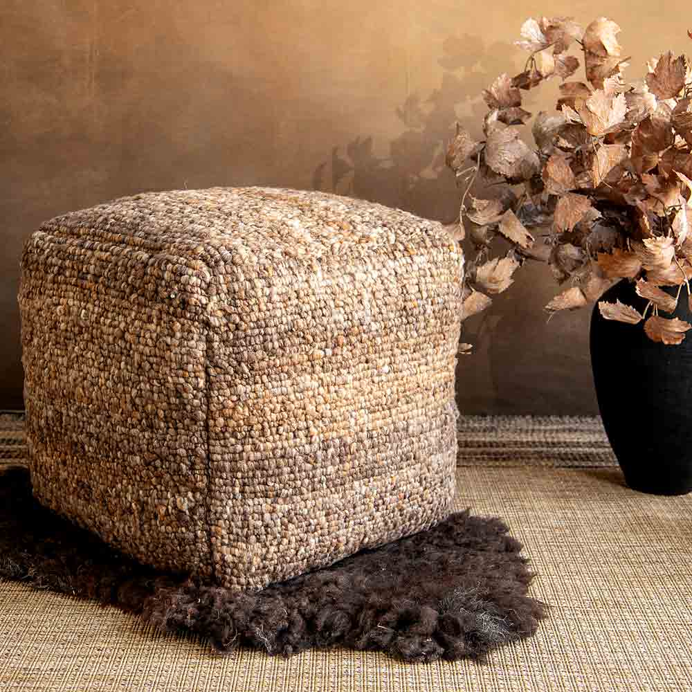 A square textured woven pouf in a beige and brown material, styled on a dark brown sheepskin rug next to a black vase with faux vine leaves against a warm brown wall.