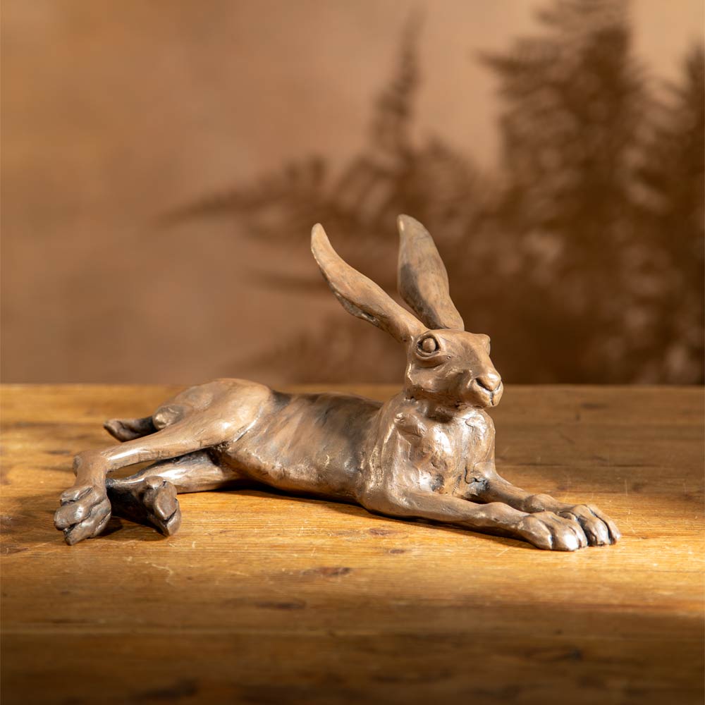 Bronze-finished sculpture of a hare lying down, displayed on a rustic wooden table against a light gray wall with a soft shadow cast behind it.