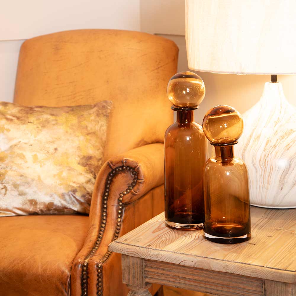 Two Lana amber glass bottles with Ball Tops (Large and Small Sizes) on a wooden table with a textured chair and lamp in the background.
