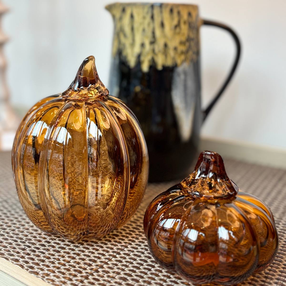 Two glass pumpkins on a textured surface with a blurred pitcher in the background.