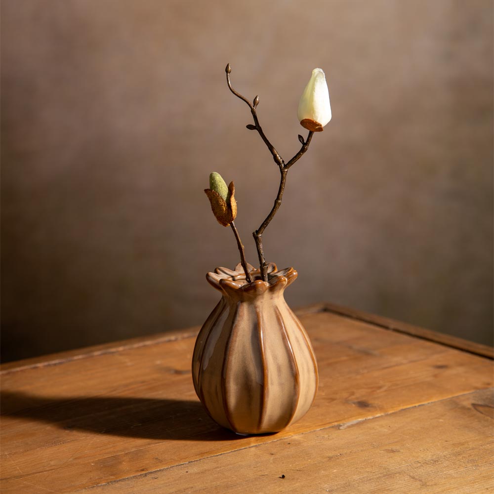 A small ceramic bud vase with a flared rim sits on a wooden table against a warm brown wall. The vase is a warm taupe and brown colour with subtle vertical ribbing.