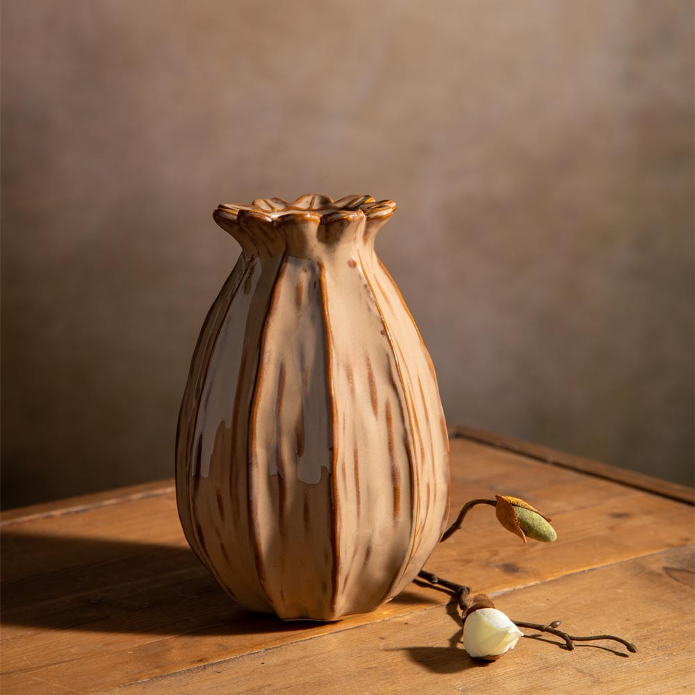 A ceramic bud vase with a glossy brown and beige glazed finish, styled on a rustic wooden table with a faux flower stem against a textured brown background.