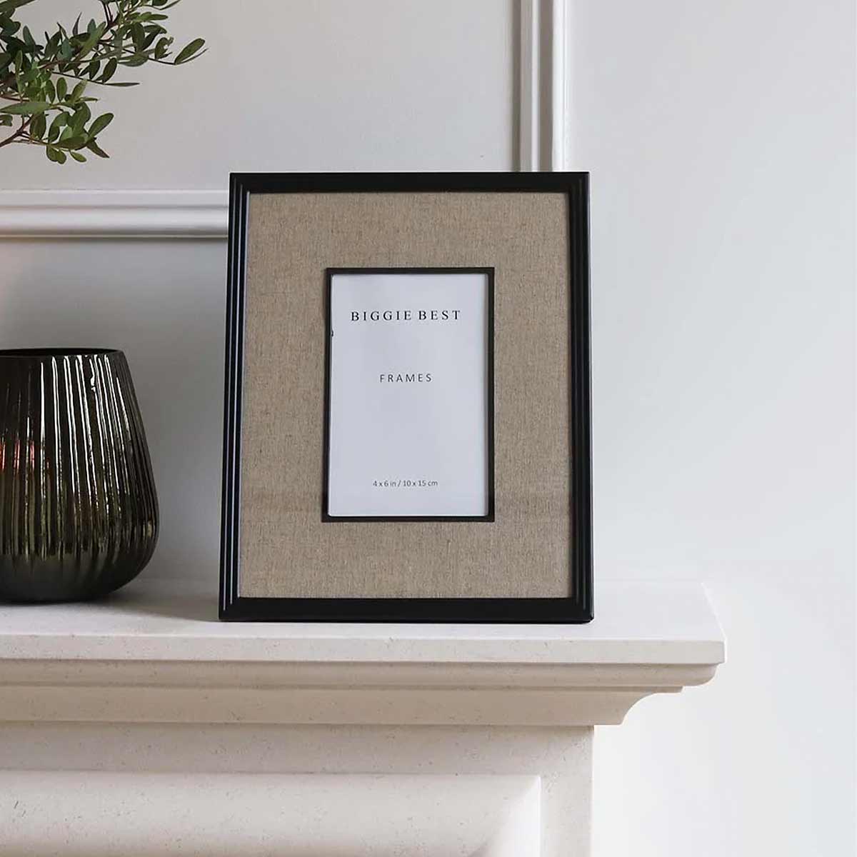 A black photo frame with a linen border displaying - 4×6 inch size. The frame is placed on a white mantelpiece with a vase and greenery.