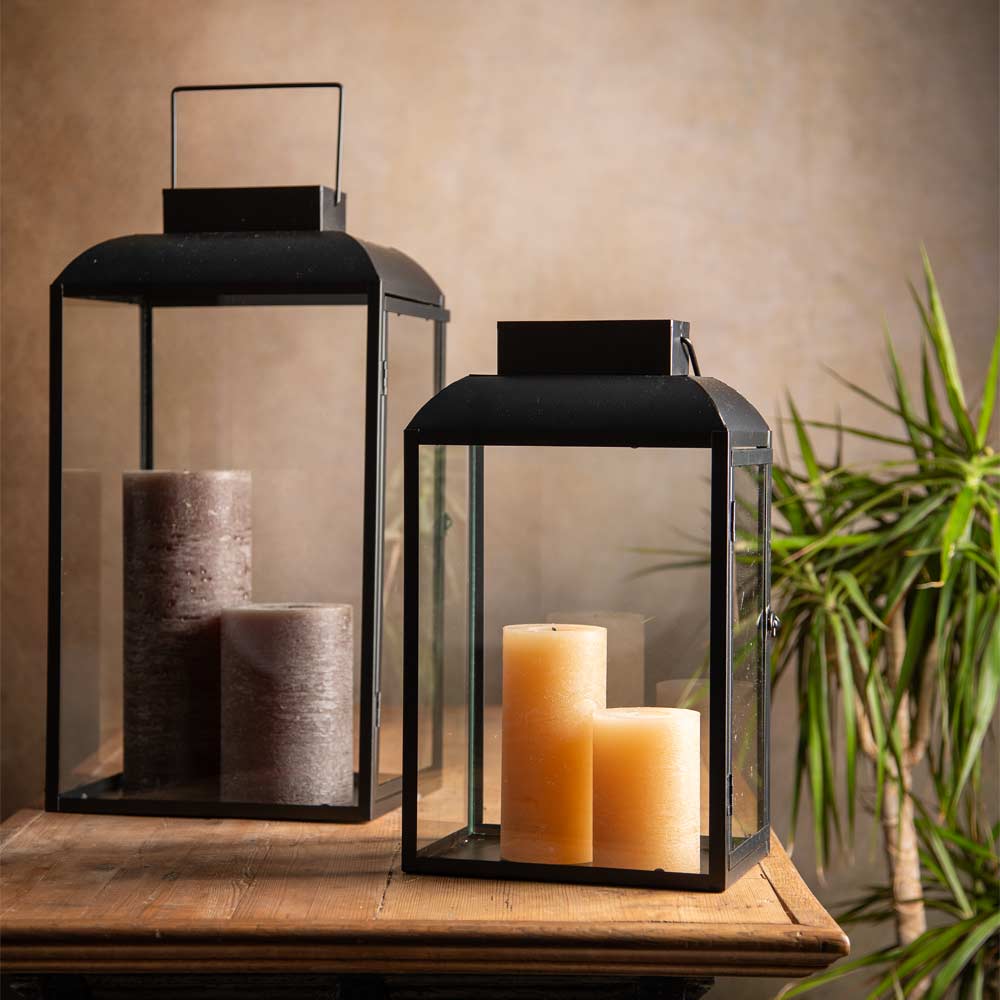 Two black metal lanterns with clear glass panels, one larger with two dark pillar candles and one smaller with two honey coloured pillar candles, displayed on a wooden table against a warm beige wall with a green plant in the background.
