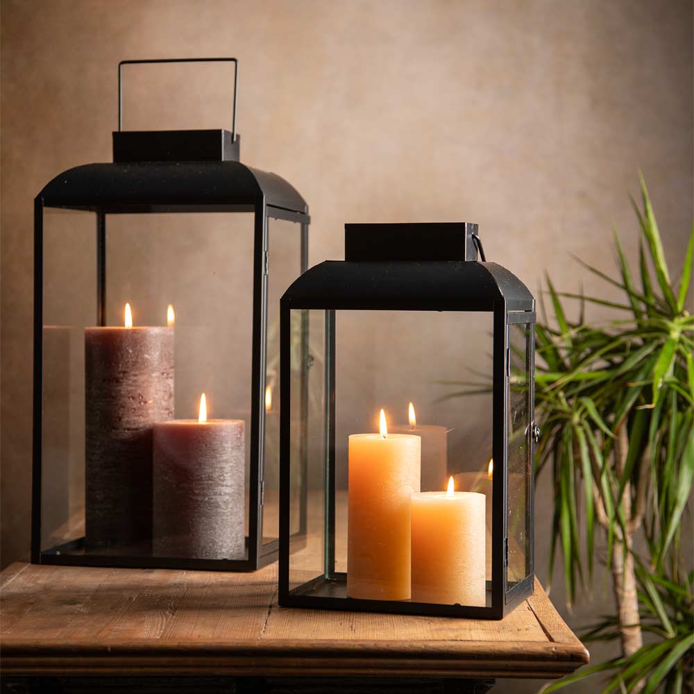 Two black metal lanterns with clear glass panels, each containing lit pillar candles, displayed on a wooden surface against a warm beige wall with a green plant in the background.