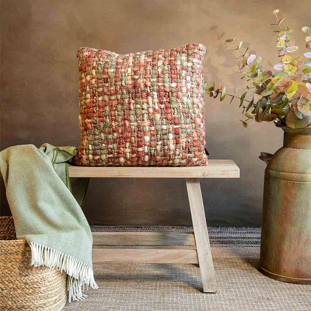 A square woven throw cushion in a textured blend of rust red, beige, and muted green sits on a light wood bench. The scene is styled with a green throw blanket draped over a woven basket on the left and a rustic metal vase with faux flowers on the right, set against a warm brown wall.