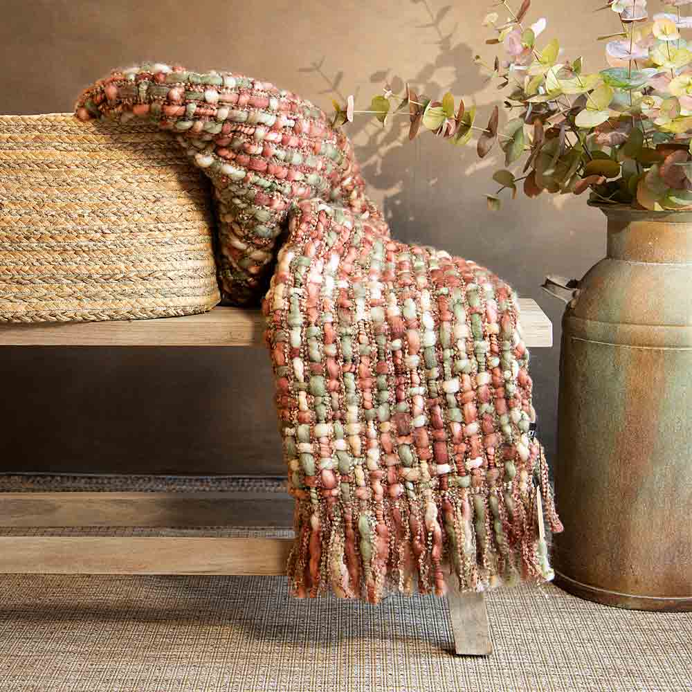A chunky woven throw blanket in a muted multicolor mix of terracotta, sage green, cream, and rust, draped over a wooden bench next to a woven basket and a rustic metal vase with autumnal eucalyptus.