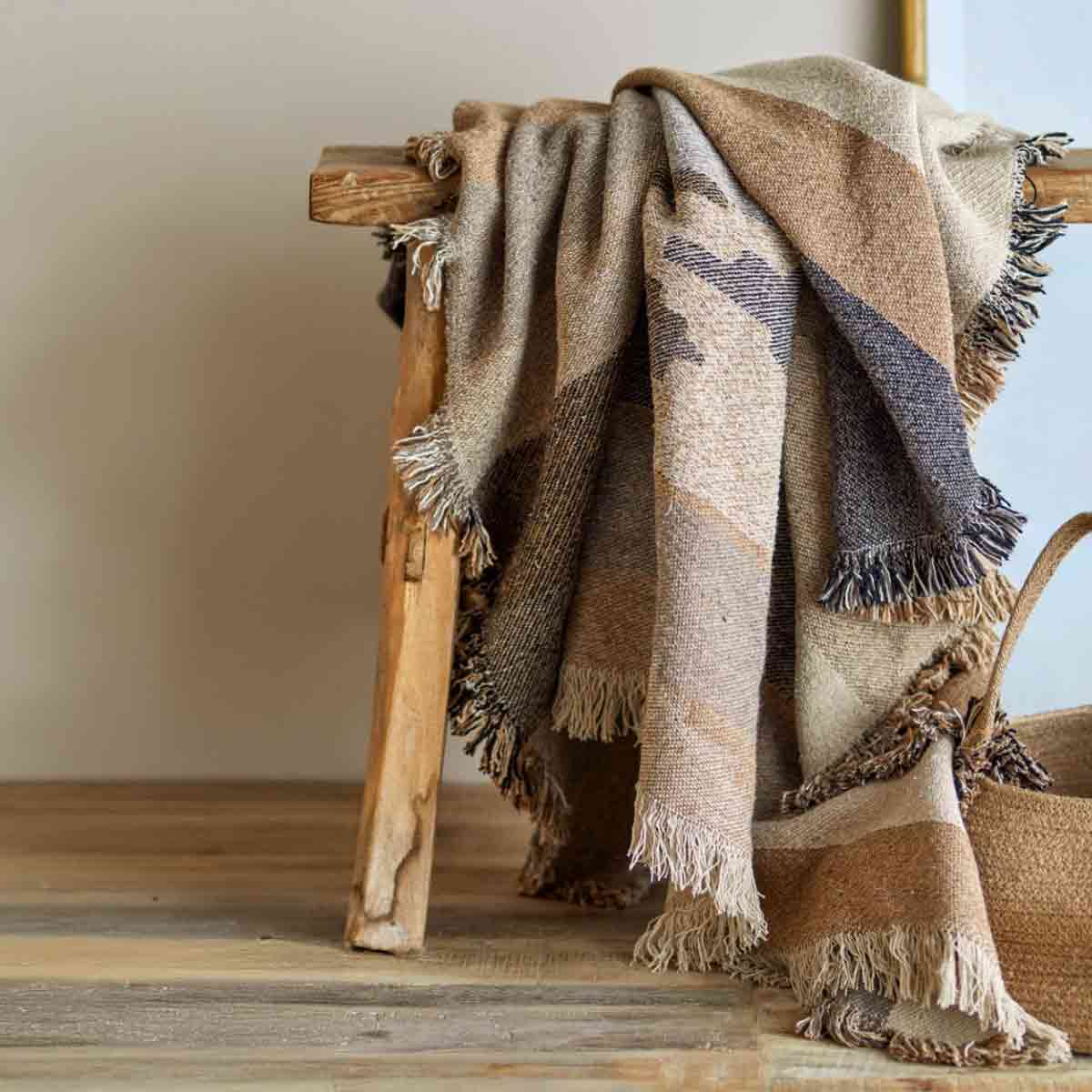 Maji Throw – a woven recycled cotton blanket in warm brown and beige tones with a subtle geometric pattern and fringed edges, draped over a rustic wooden stool.