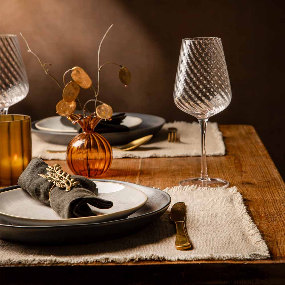 A styled dining table setting with clear textured wine glasses, layered charcoal grey and white plates, beige placemats, gold-toned cutlery, and a small amber glass vase with faux leaves on a wooden table.