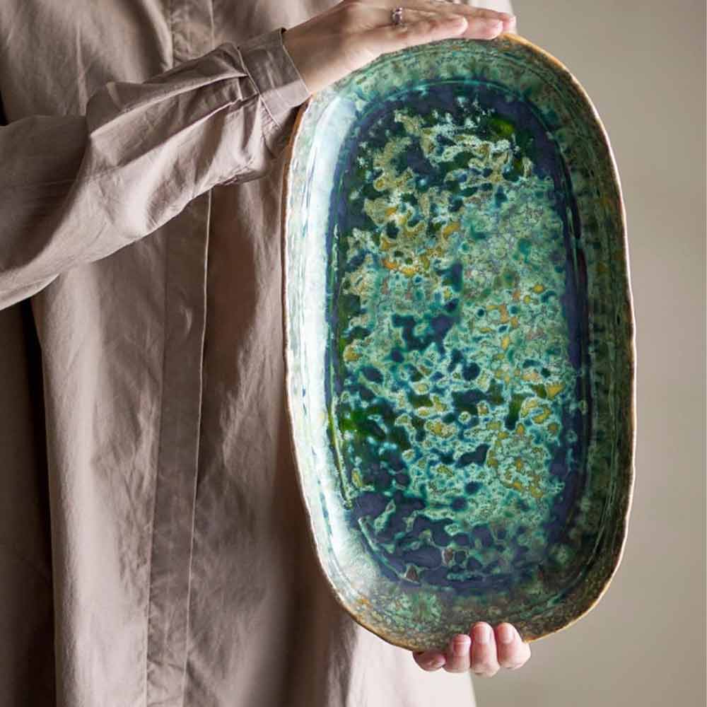 Hands holding the Milani Green Stoneware Serving Plate, showcasing its reactive glaze with deep green, ocean blue, and golden hues.