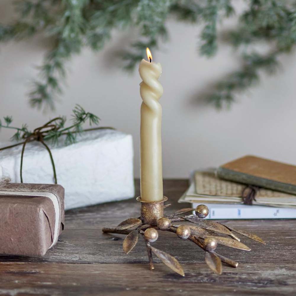 Mirabel Gold Candle Holder with mistletoe leaves and berries with lit taper candle on a blurred background of books and greenery