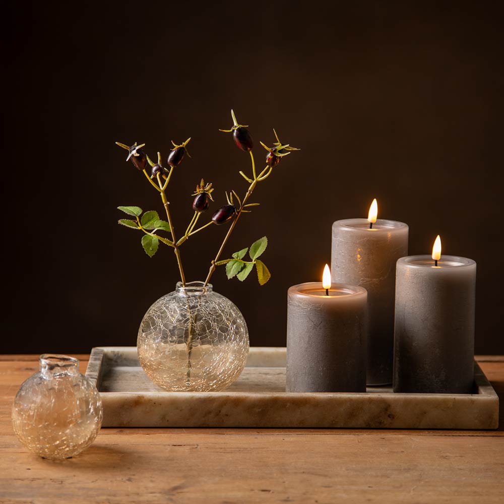 A styled tabletop arrangement with three lit mocca pillar candles on a marble tray beside a small clear crackled glass vase holding faux stems, and another small crackled glass vase on the left, against a dark brown background.