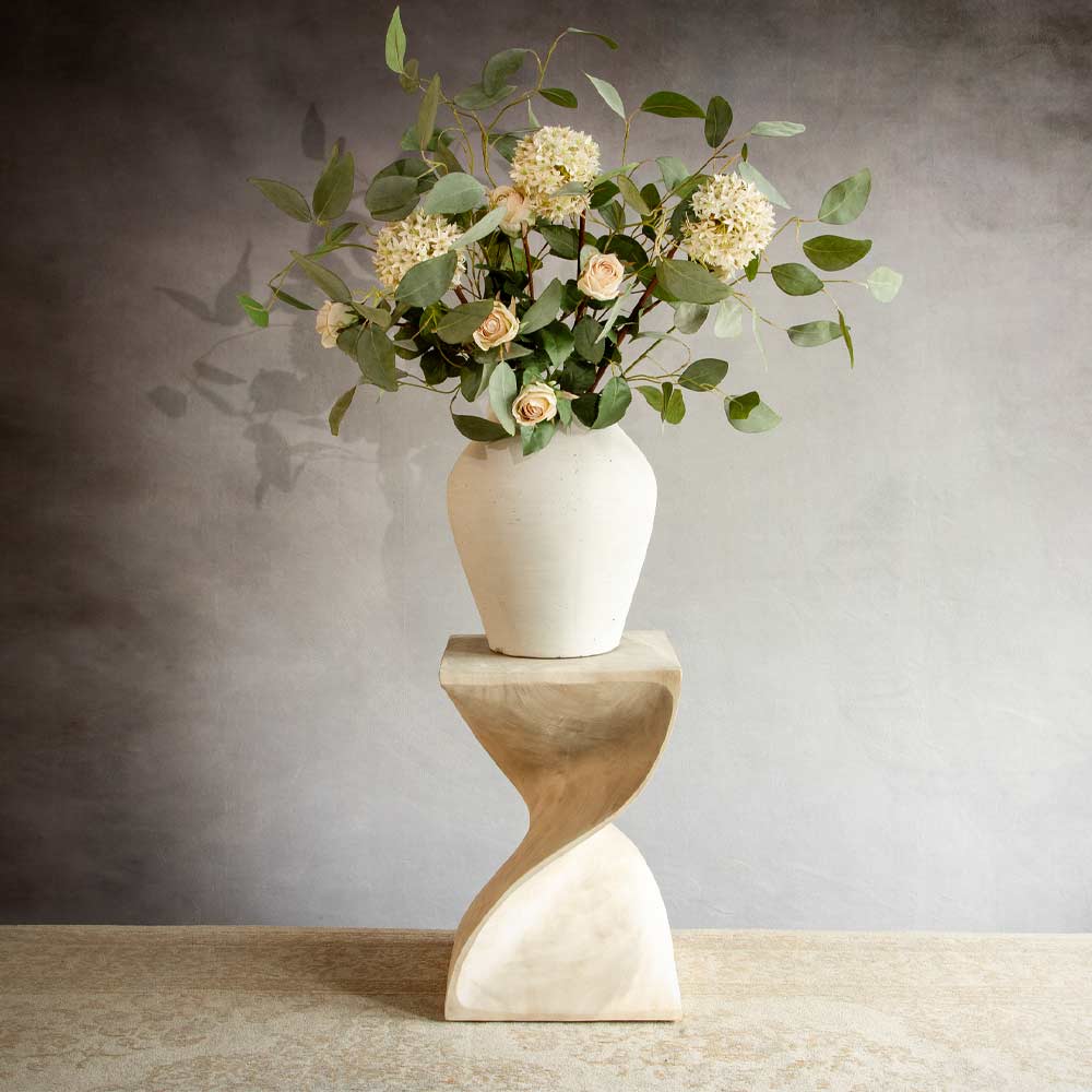 A cream ceramic vase filled with green eucalyptus leaves, cream roses coloured viburnum flowers, displayed on a sculptural twisted wood pedestal against a textured grey wall.