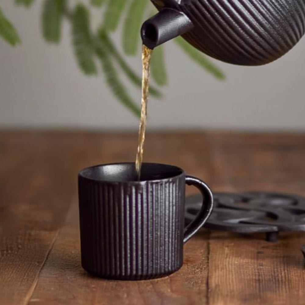 Black teapot pouring tea into a Neri Stoneware Mug, Black on a wooden surface