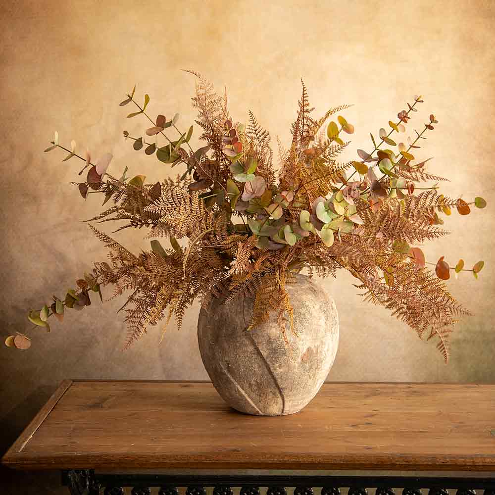 A faux floral arrangement with ferns, eucalyptus leaves, in a round textured beige and brown rustic textured ceramic vase, displayed on a wooden table against a warm beige wall.