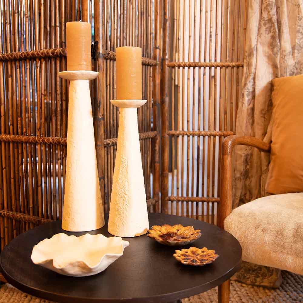 Decorative table setting with Ota Cream Pillar Candle Holders in two sizes styled with candles and bowls against a woven wall and chair.