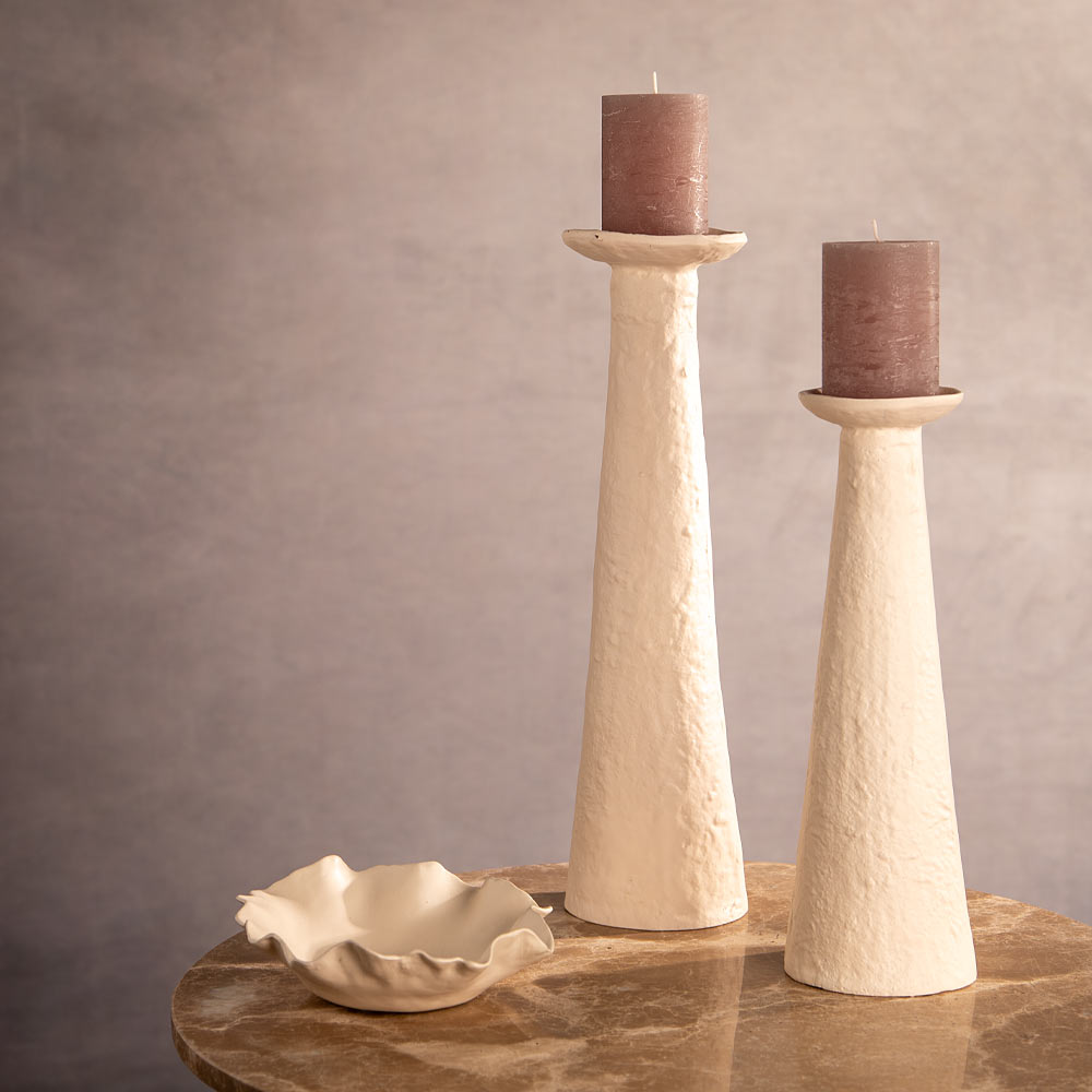 Decorative table setting with Ota Cream Pillar Candle Holders in two sizes styled with candles and bowls against a woven wall and chair.