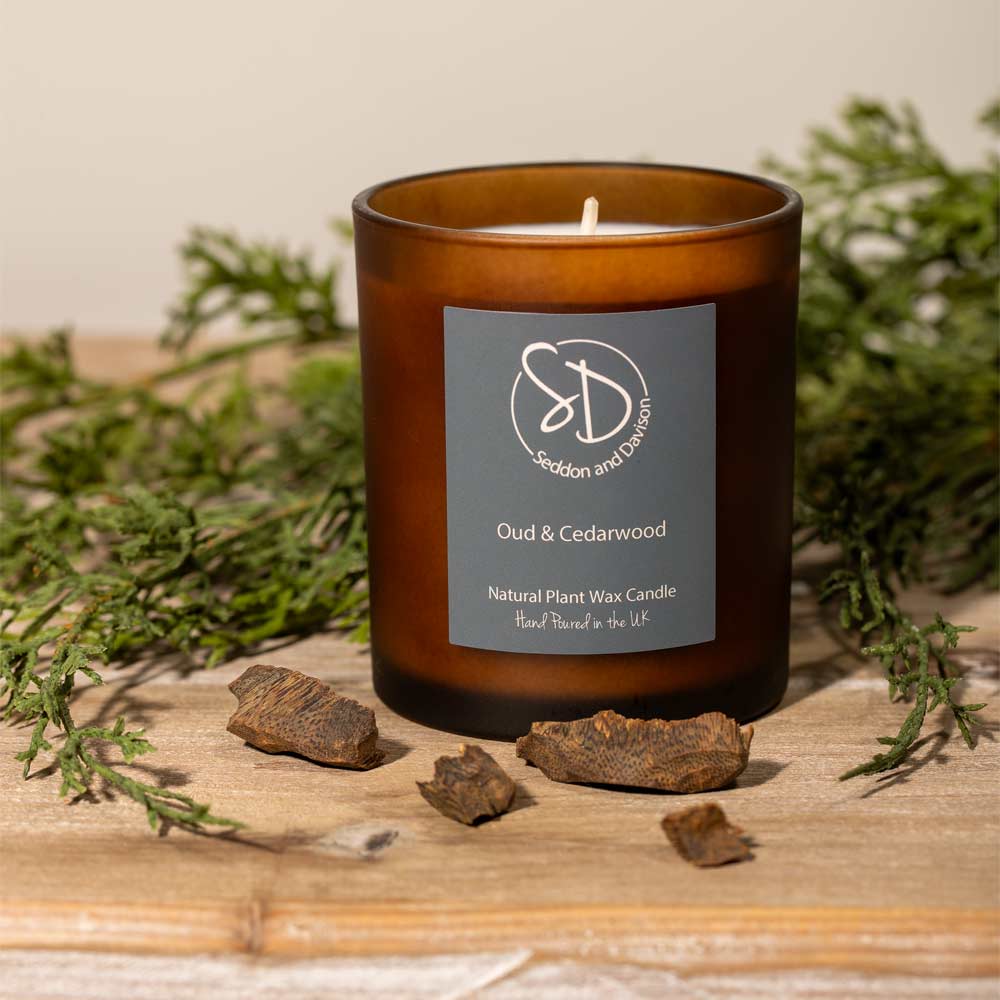 An amber glass jar candle with a gray label reading 'Oud & Cedarwood' and the 'Seddon and Davison' logo, styled on a wooden surface with green foliage and dried bark pieces.