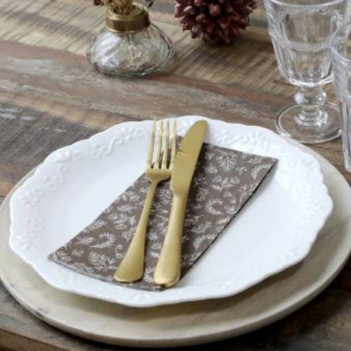 Set table with white plates, gold cutlery, and a brown patterned paper napkin with white leaf patter on a wooden rustic table.