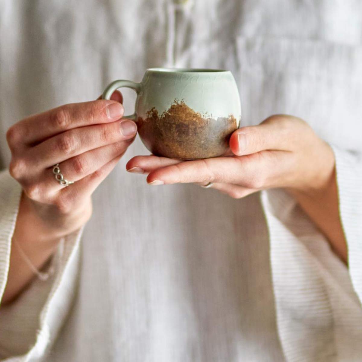Paula Stoneware Espresso Cup in brown and blue-green reactive glaze, featuring a rounded shape and a circular handle, held by person in a linen shirt.