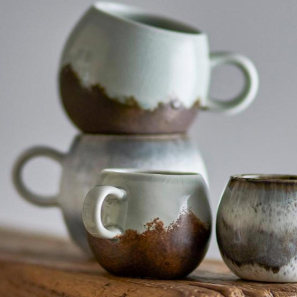 Paula Stoneware Espresso Cup in brown and blue-green reactive glaze, featuring a rounded shape and a sturdy handle, stacked with other matching cups on a rustic wooden surface.