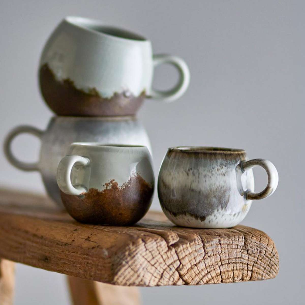 Paula Stoneware Espresso Cup in grey, white, and brown reactive glaze, stacked on rustic bench and displayed with other similar cups, showcasing its organic shape and unique colour variations.