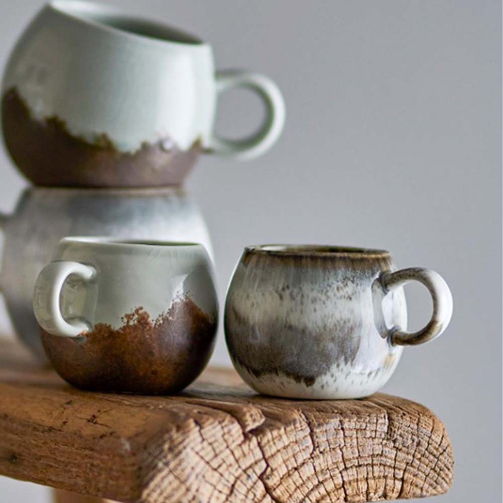 Paula Stoneware Espresso Cup in grey, white, and brown reactive glaze, stacked on rustic bench and displayed with other similar cups, showcasing its organic shape and unique colour variations.