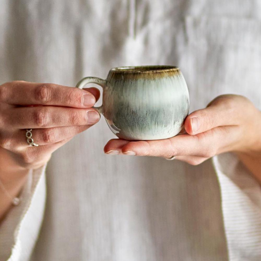 Paula Stoneware Espresso Cup in grey, white, and brown reactive glaze, held in both hands, showcasing its organic shape and unique colour variations.