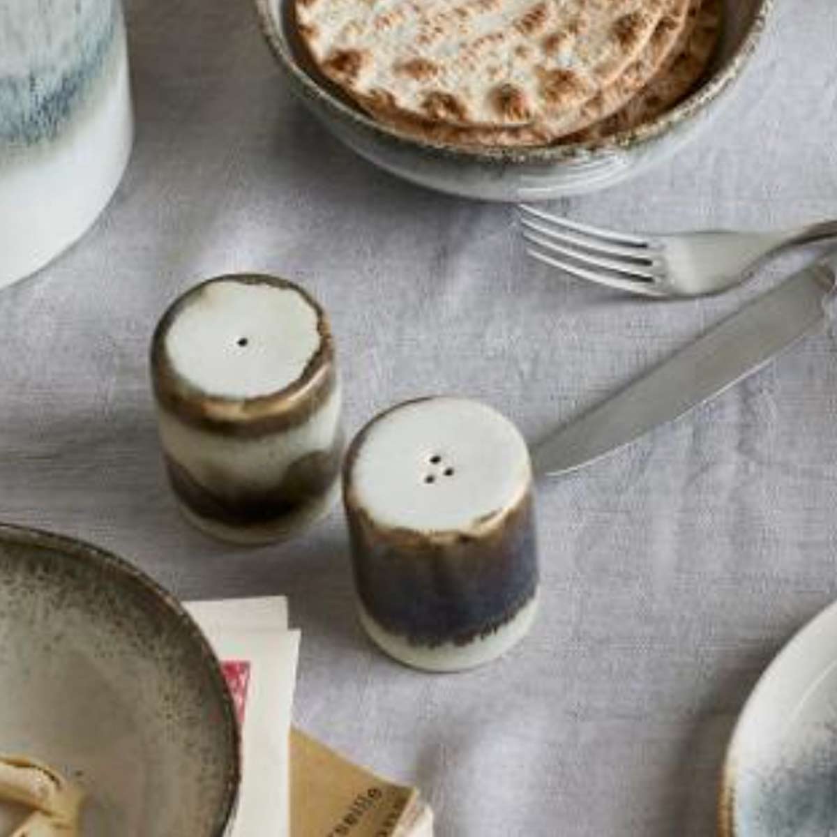 Paula Stoneware Salt and Pepper Shaker Set, Grey - A set of two stoneware salt and pepper shakers in soft grey with an organic reactive glaze, placed on a table with a linen tablecloth with bread.
