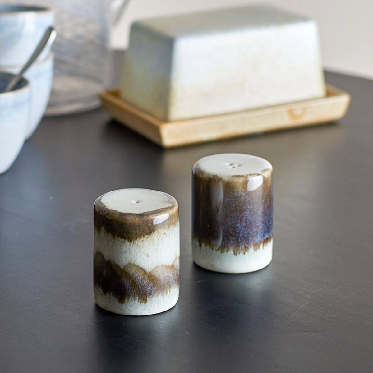 Paula Stoneware Salt and Pepper Shaker Set, Grey - A set of two stoneware salt and pepper shakers in soft grey with an organic reactive glaze, placed on a table with a textured background.
