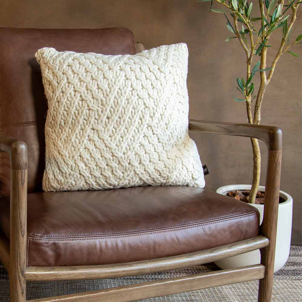 close up of a modern armchair with a brown leather seat and backrest, a light wood frame, and a cream textured knit throw pillow, styled in a rustic room setting.