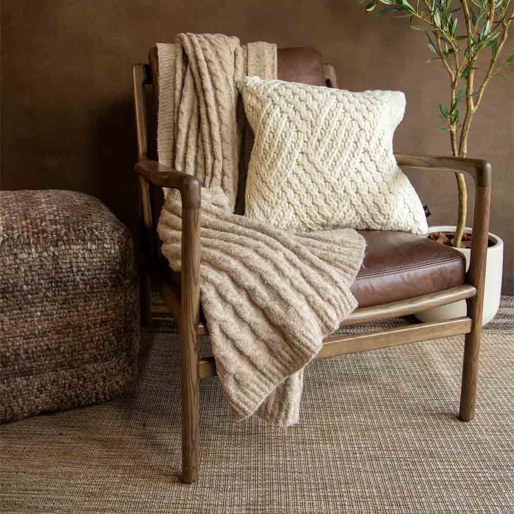 A cozy indoor scene with a modern wooden armchair featuring a brown leather seat, styled with a beige knit throw blanket and a cream textured pillow. A woven pouf sits to the left, and a potted plant is in the background against a warm brown wall.