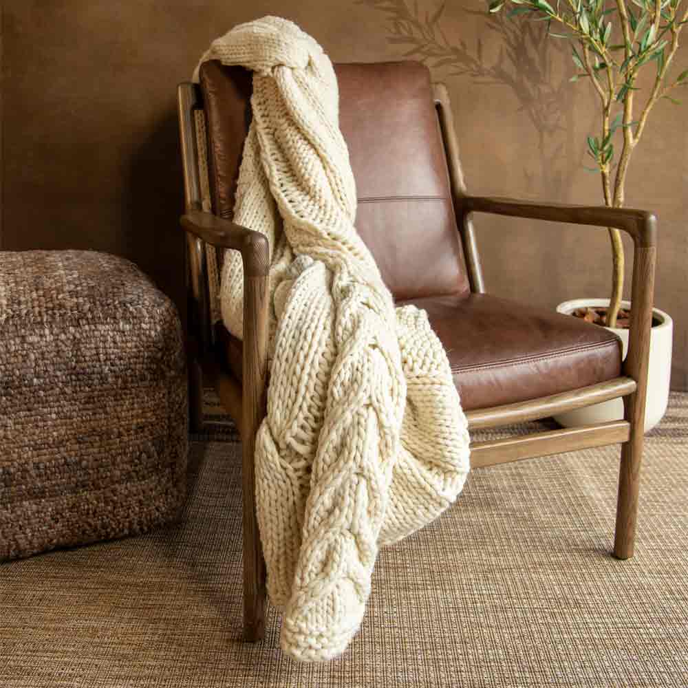 A chunky cream knit throw blanket is draped over a mid-century modern armchair with brown leather upholstery, set in a cozy room with a textured rug, a potted plant, and a woven ottoman.