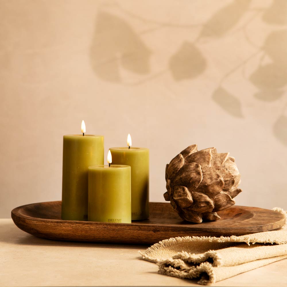Three lit LED olive green pillar candles arranged on a round wooden tray beside a decorative brown artichoke, with a folded beige fabric in the foreground against a warm beige wall with soft leaf shadows.