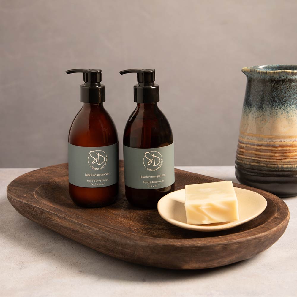 Two amber pump bottles with muted grey labels reading Black Pomegranate Hand & Body wash and Black Pomegranate hand & Body Lotion , styled on a dark mango wood tray beside a small dish with soap bar and a ceramic pitcher against a neutral gray background.