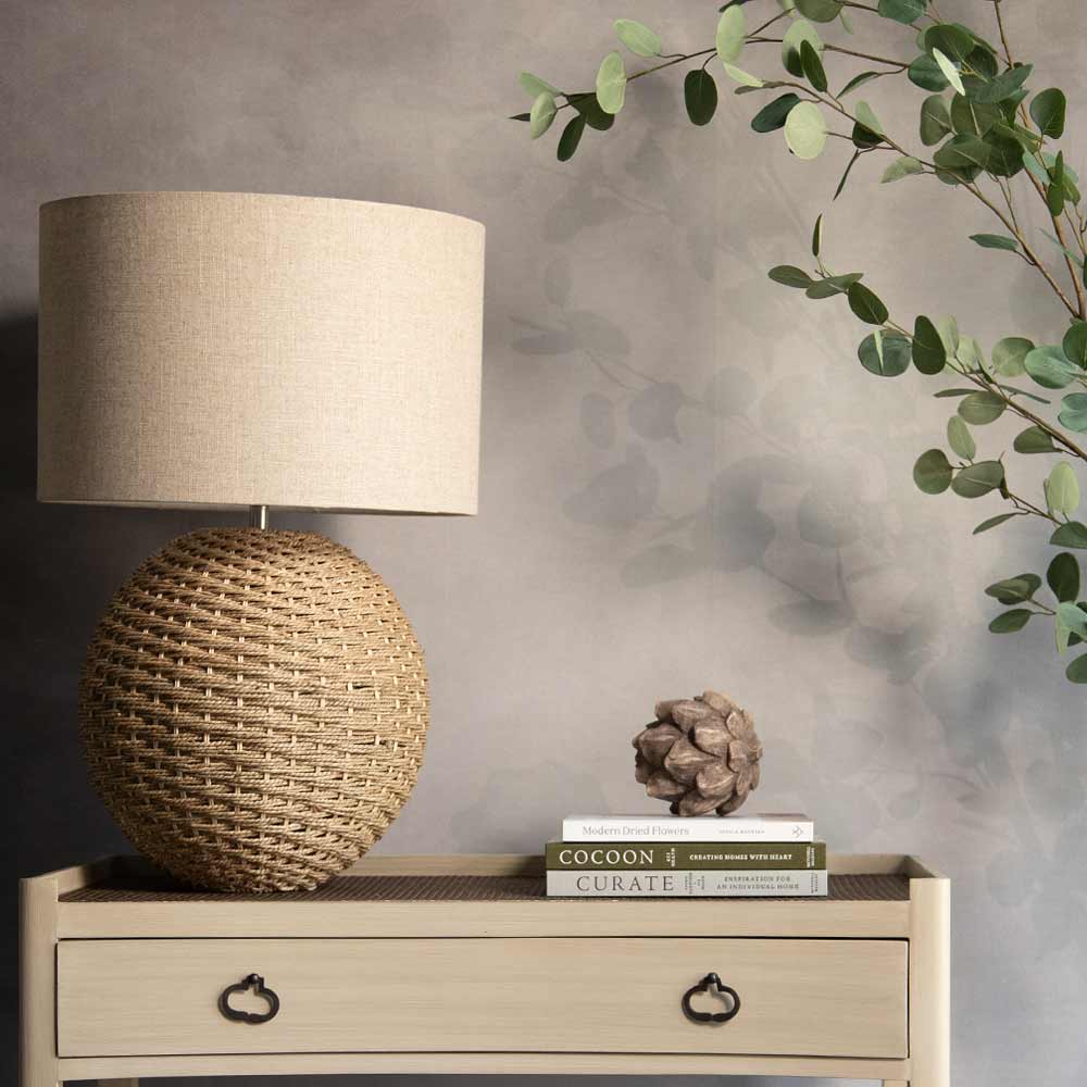 A woven spherical table lamp with a beige drum shade sits on a light wood console table against a textured gray wall, styled with books and a decorative object, with eucalyptus branches in the upper right.