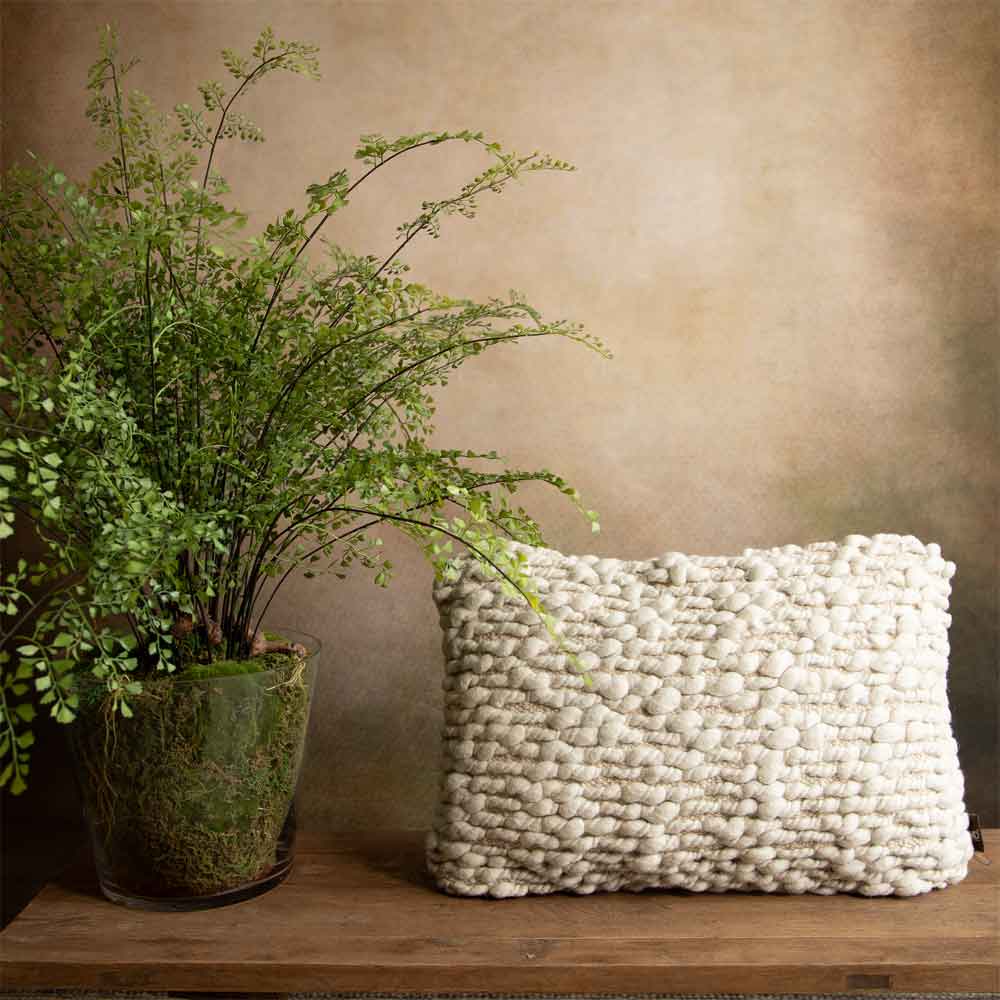 A oblong , chunky-knit throw pillow in a warm off-white/cream colour, styled on a wooden surface next to a potted green fern against a textured beige wall.