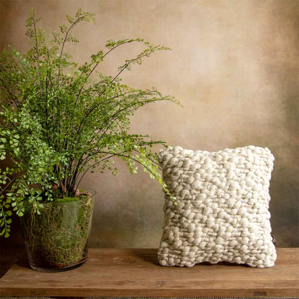 A square cushion with a chunky, textured surface in an off-white cream color, styled on a wooden bench next to a potted green fern against a warm, textured beige wall.