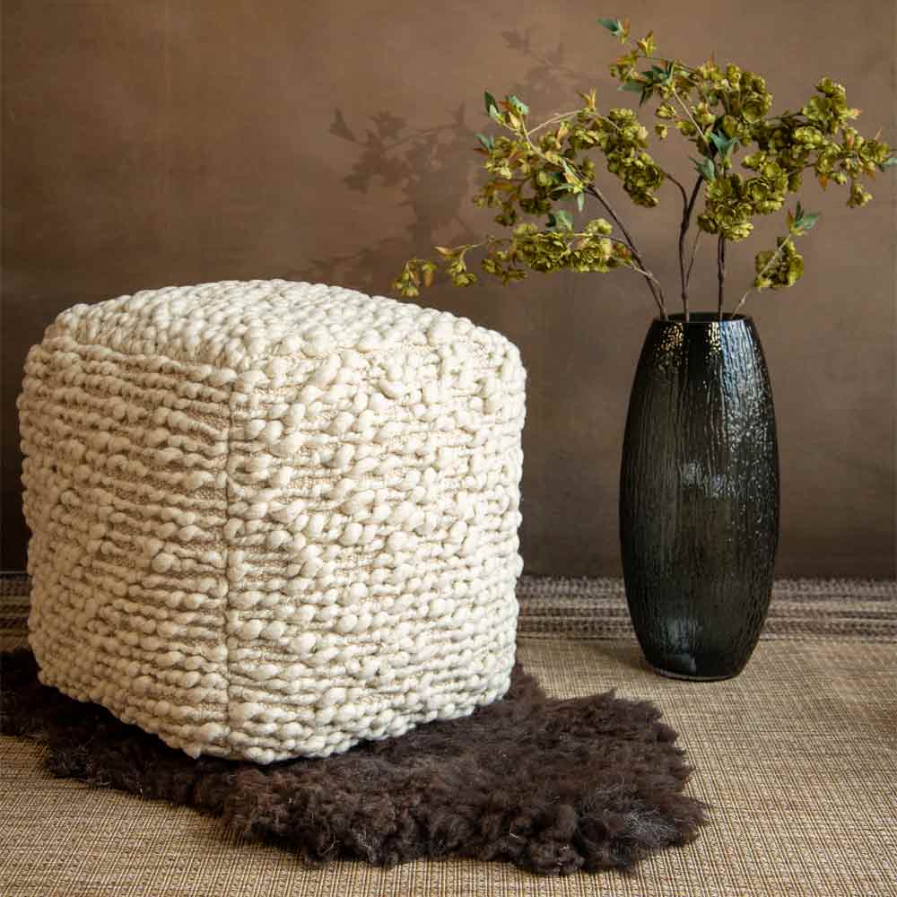 A chunky cube pouf in cream-coloured yarn, styled on a dark brown sheepskin rug next to a black textured glass vase with dried green foliage, set against a warm brown background.