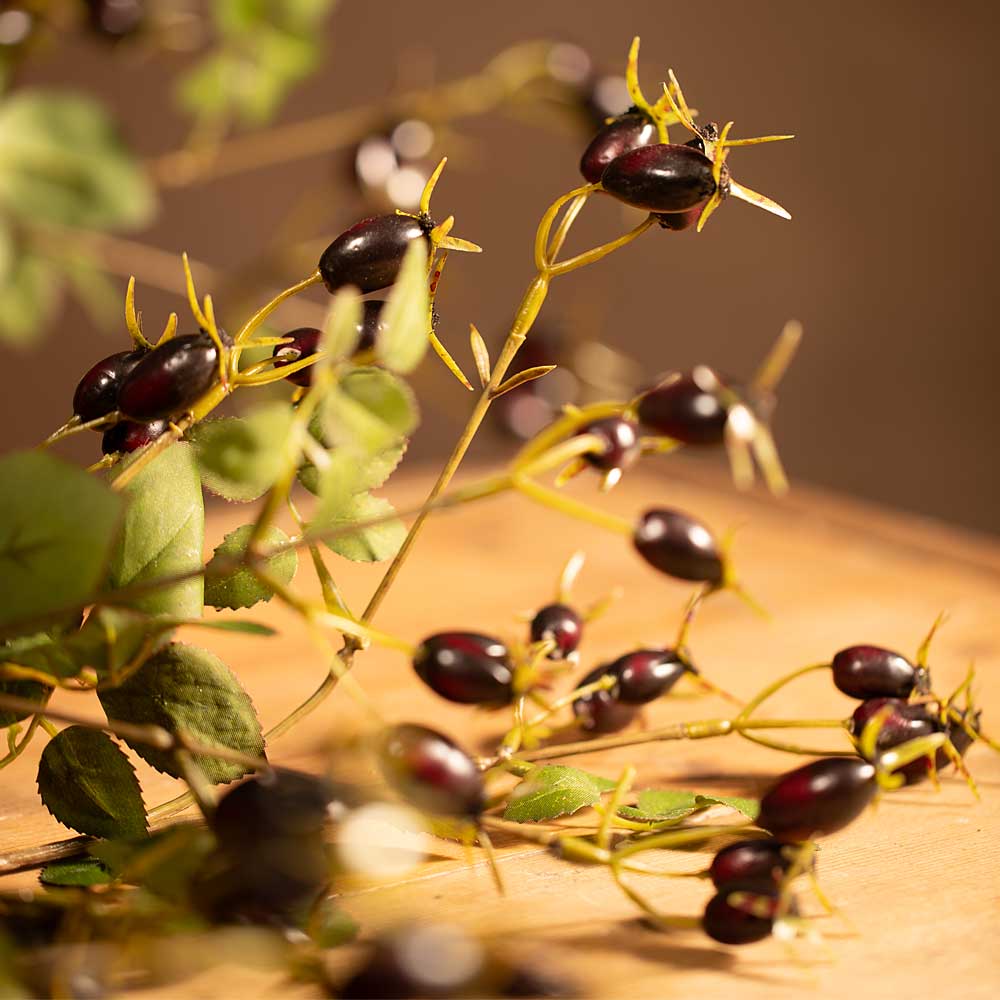 Faux rose hip branch with green leaves, displayed on a wooden surface with a warm, soft-focus background.