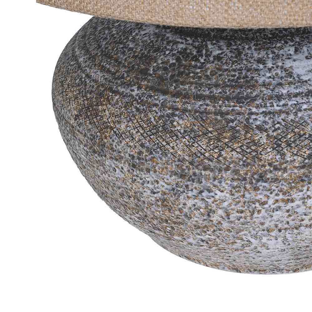 Close-up of a decorative ceramic table base with a textured, speckled pattern in gray, white, and rust, topped with a woven style beige fabric shade on a white background.