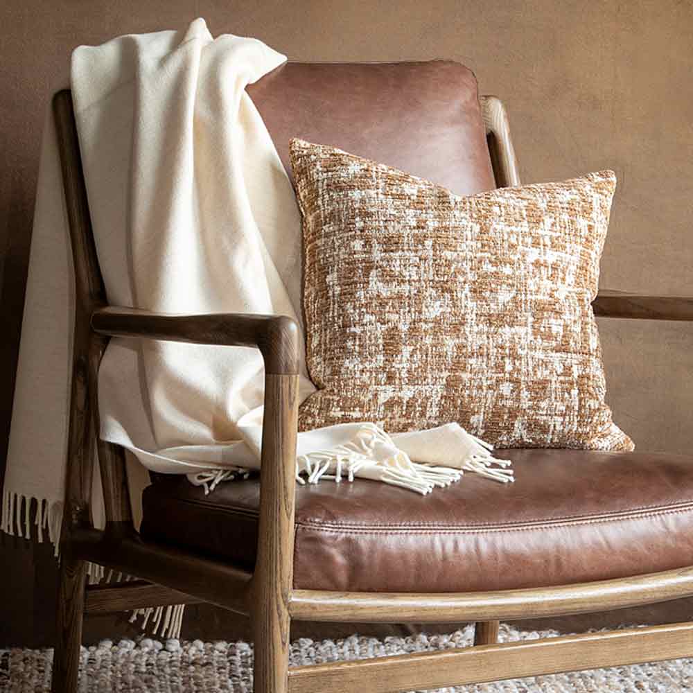 A brown leather armchair styled with a cream throw blanket draped over the side and a textured cream and brown textured pillow resting on the seat, set against a warm brown wall.